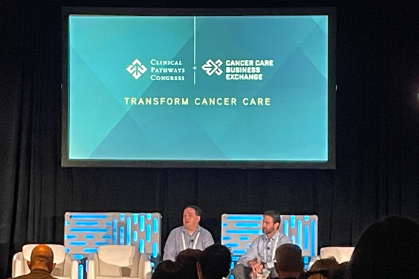 Two speakers sitting in chairs presenting at CB Exchange. Presentation slide shows two logos: Clinical Pathways Congress (on left) and Cancer Care Business Exchange (on right) as a logo mash up. Under the logos, there is the slogan, "Transform Cancer Care"