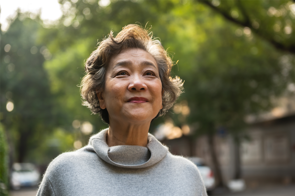 Older woman standing outdoors. She is wearing a light gray turtle neck